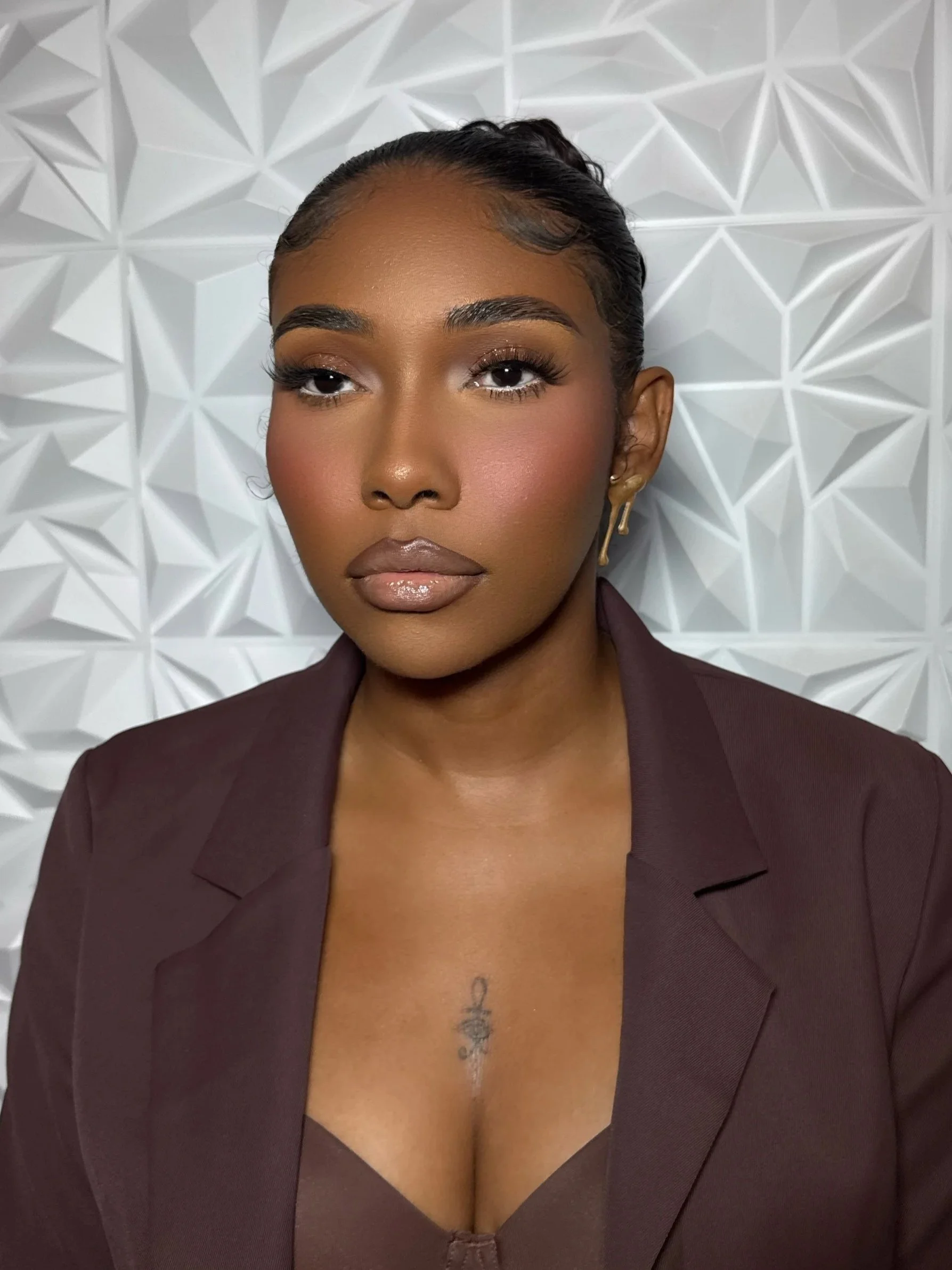 A woman with her hair slicked back, wearing makeup, gold earrings, and a dark blazer, against a white textured geometric wall.