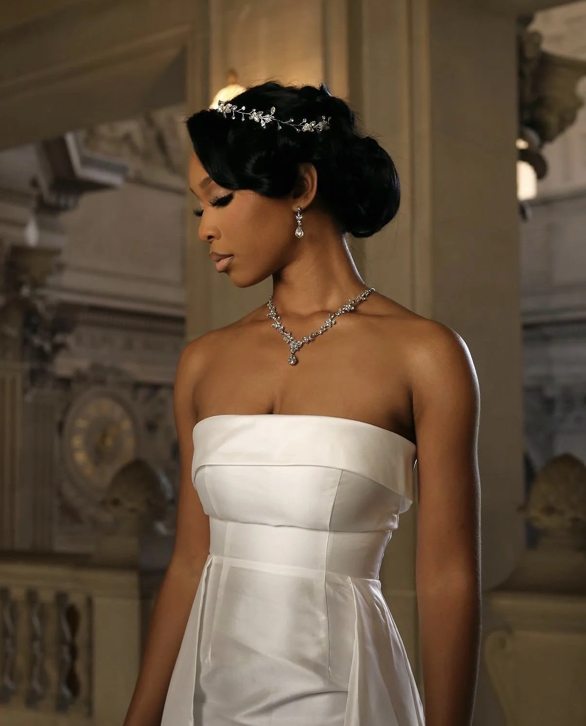A woman in an elegant white strapless gown with jewelry, standing in a grand ornate interior.