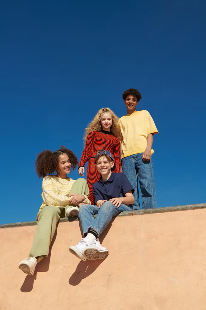 Four kids smiling and sitting on a skate park ledge against a clear blue sky, wearing colorful casual clothing.