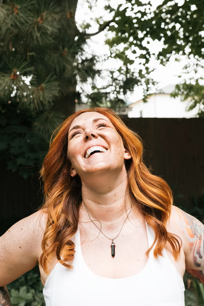 A woman with long red hair smiling and looking upward outdoors, with trees and a house in the background.