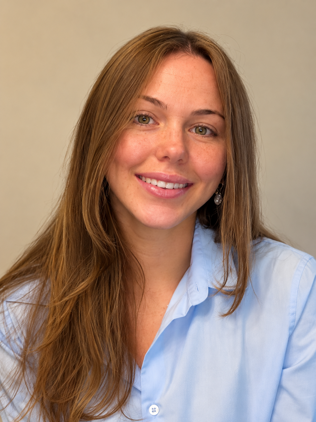 A smiling woman with long, wavy brown hair wearing a light blue button-up shirt and earrings, against a plain beige background.