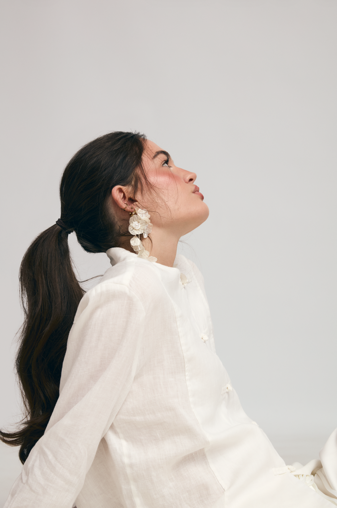 A woman with dark hair tied back, wearing a white top, large floral earrings, looking sideways against a plain background.