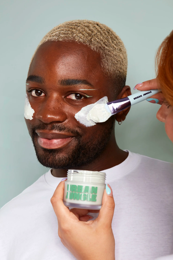 Person with blonde curly hair getting face moisturizer applied with a brush, holding a container labeled "Urban Jungle".