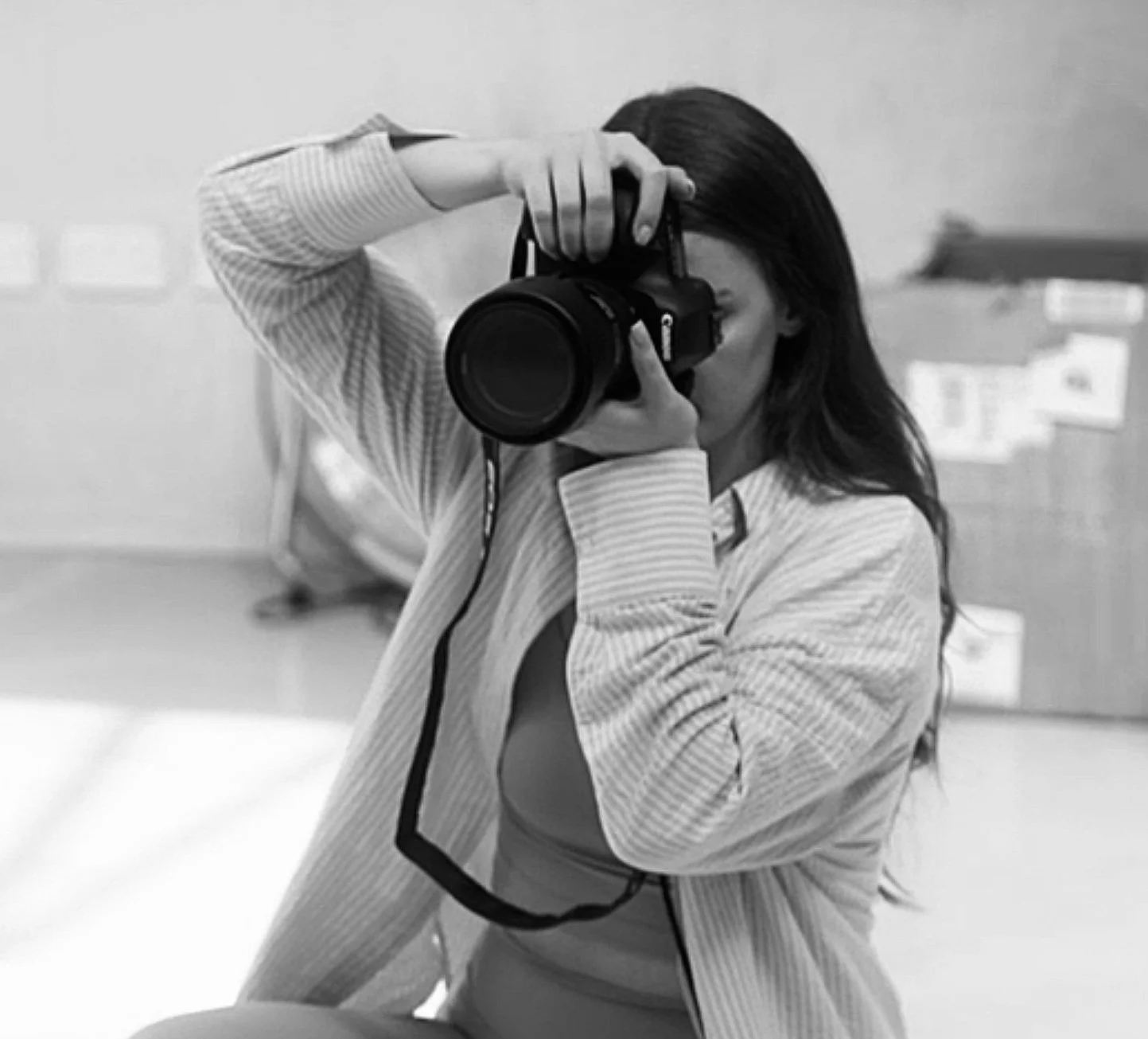 A woman with long dark hair taking a photo with a professional camera in an indoor setting, wearing a light-colored, striped jacket.