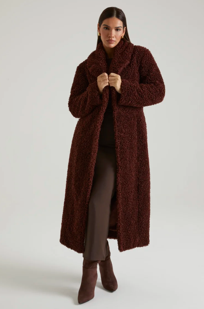 A woman standing against a plain background, wearing a long, burgundy, fuzzy coat over dark pants and boots, with her hands holding the collar of the coat.