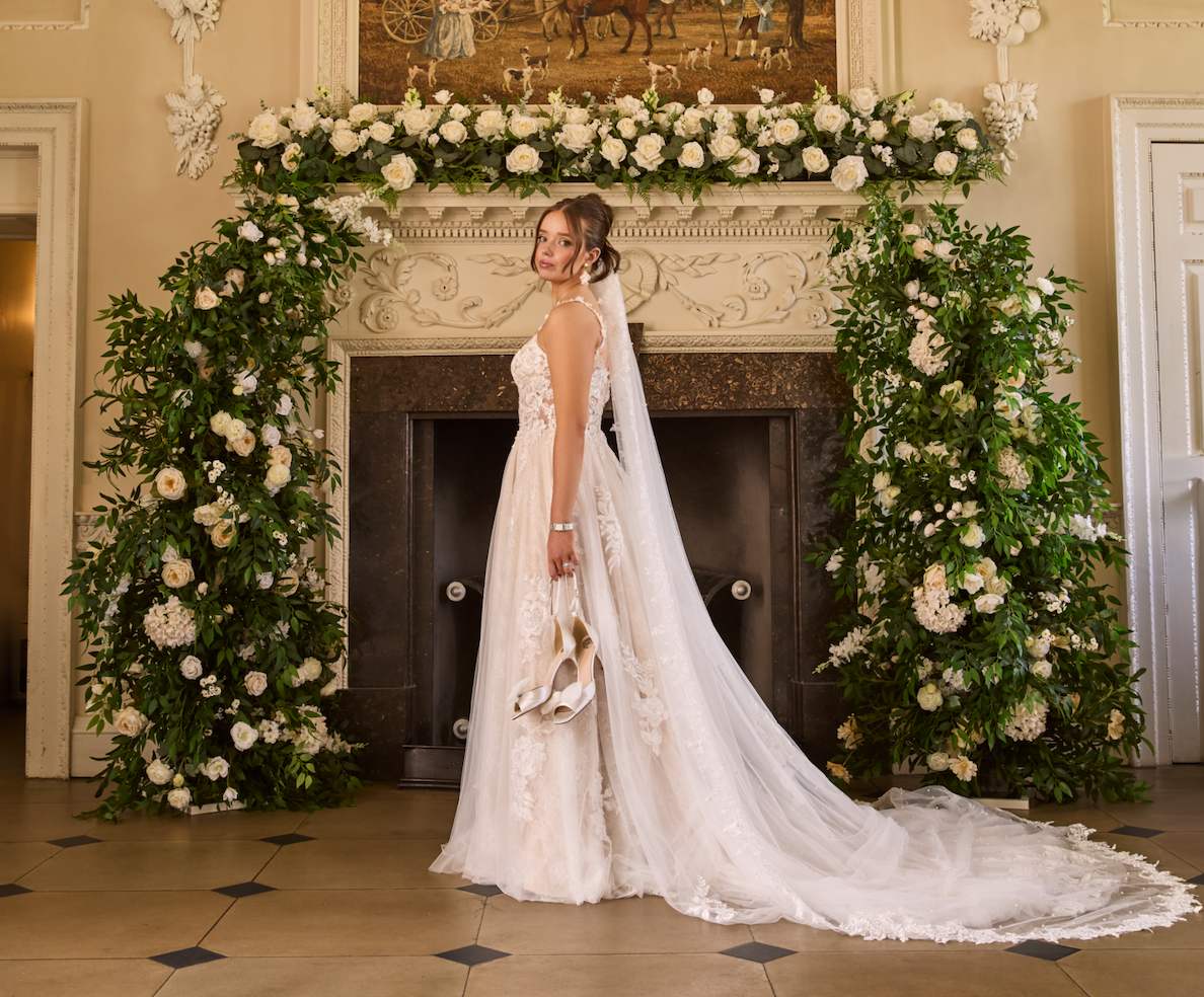 A bride in a wedding gown holding her shoes stands in front of a decorative fireplace surrounded by an arch of white flowers and greenery, with a framed painting above.