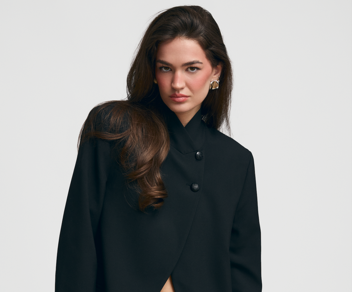 A young woman with long brown hair wearing a black blazer and gold earrings, posing against a plain light background.