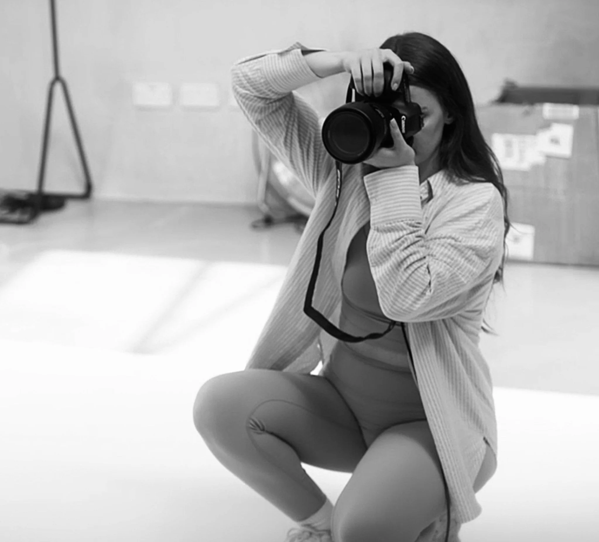 A woman with long dark hair wearing a striped blazer, taking a photo with a DSLR camera in a modern indoor space.