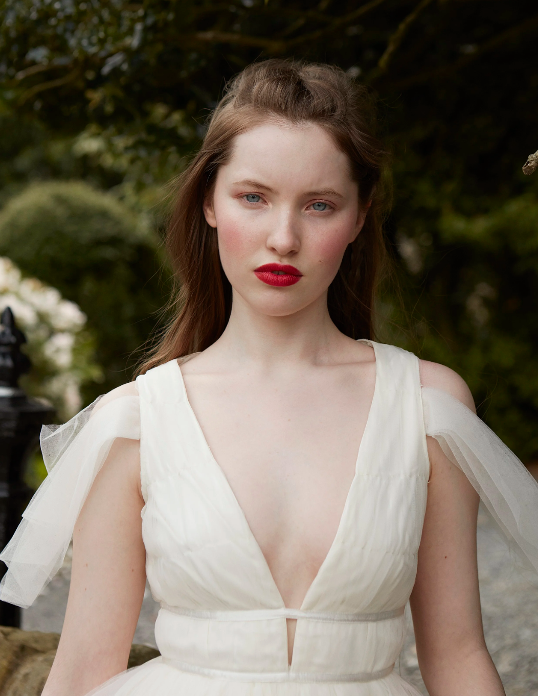A young woman with light skin and red hair, wearing a white dress with tulle accents on the shoulders, standing outdoors with greenery in the background.