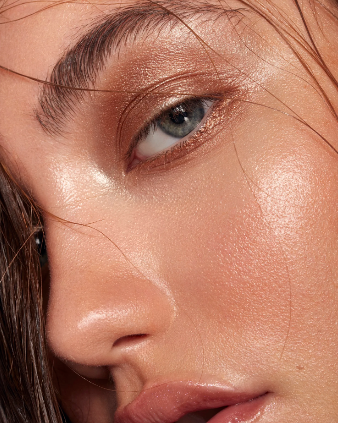 Close-up of a woman's face showing her eye, eyebrow, nose, and lips with makeup and glowing skin.