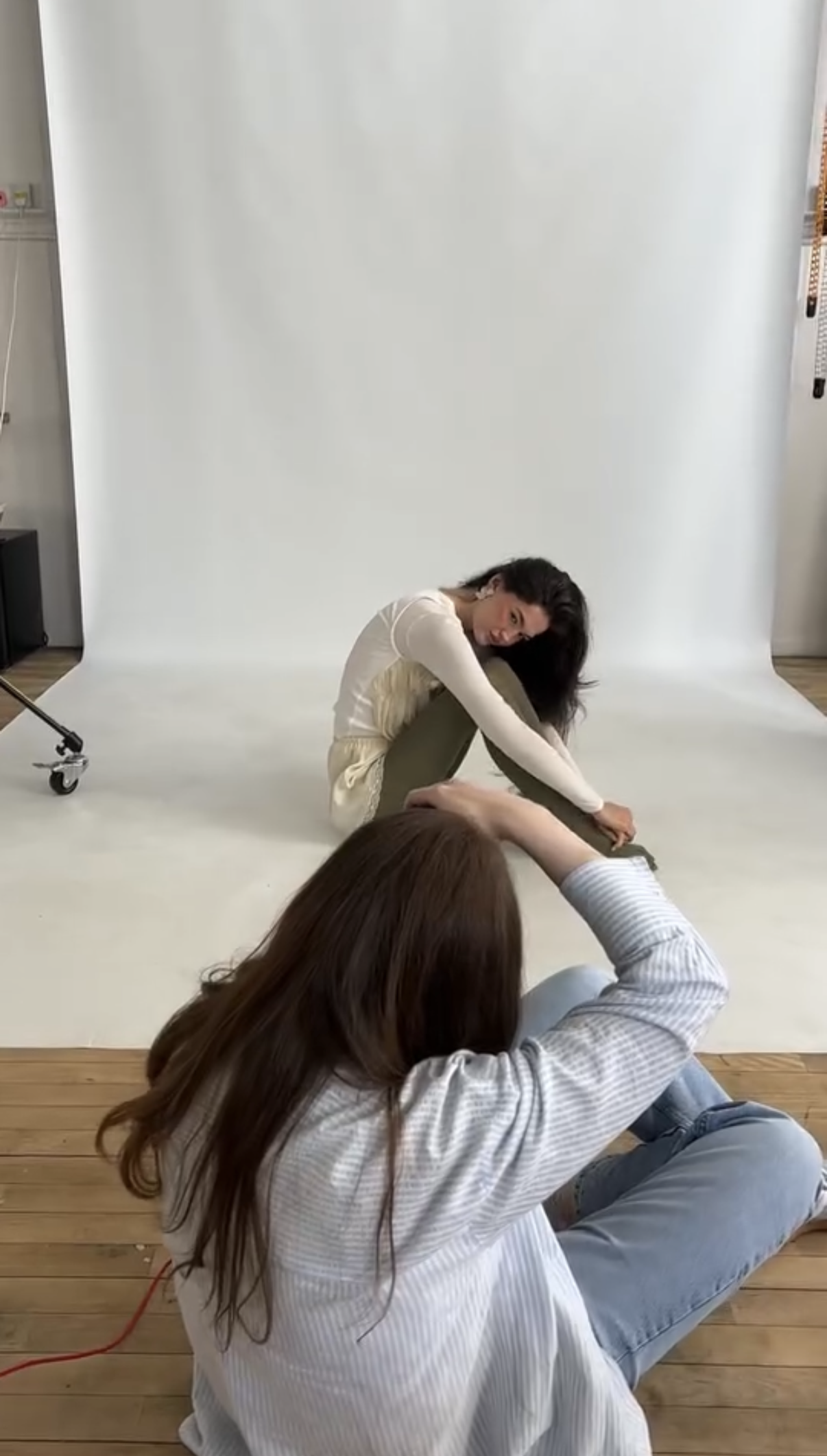 A photoshoot setup with a woman sitting on a white backdrop, posing with her legs crossed. A photographer is lying on the wooden floor taking her picture, while another person is sitting near her, partially visible.