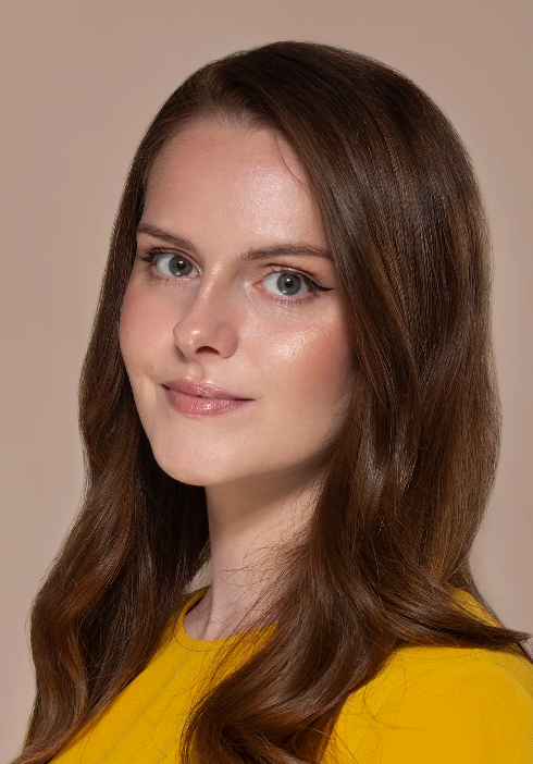 A woman with long brown hair, blue eyes, and light skin wearing a yellow top, looking at the camera with a slight smile.