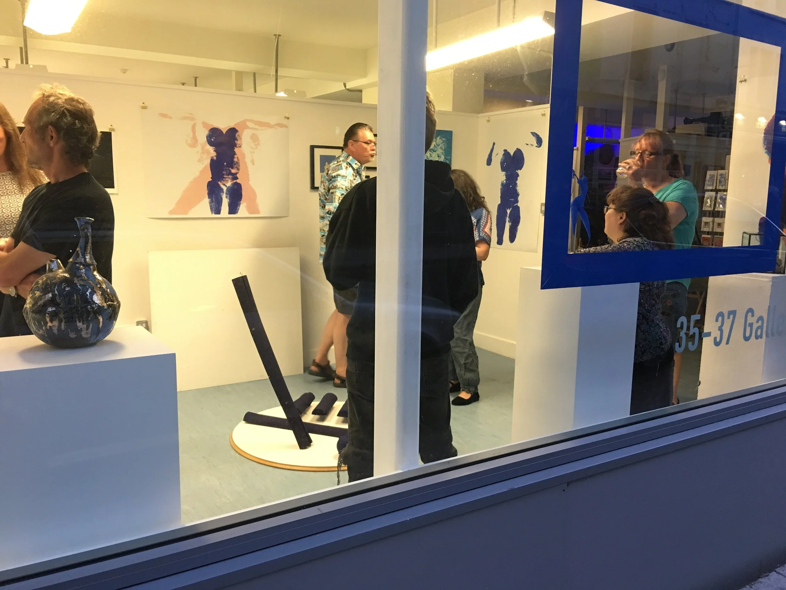 An image showing an art exhibition through a window with people looking at the artworks