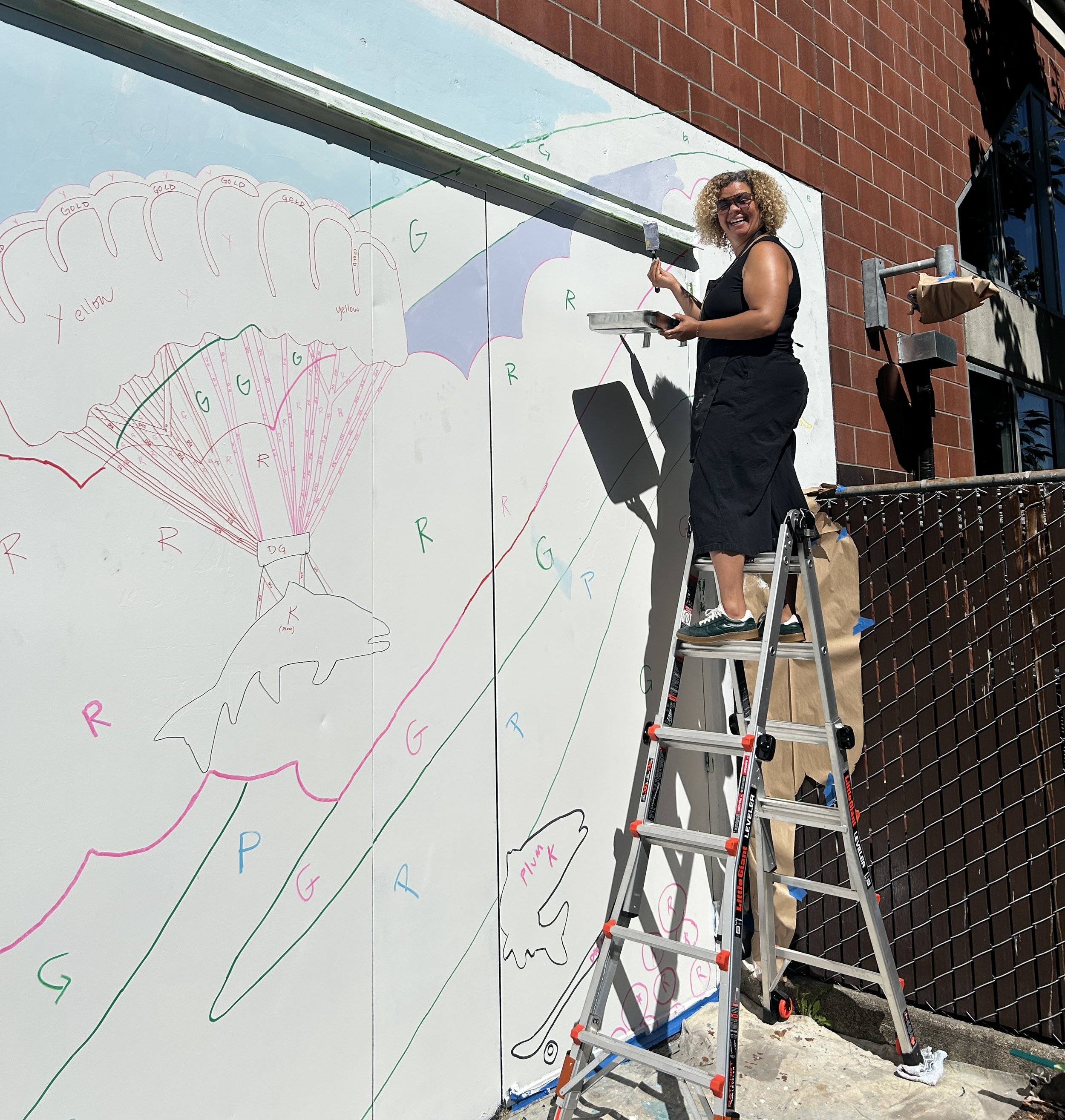 Sarah is on a ladder with a paintbrush helping to paint a mural