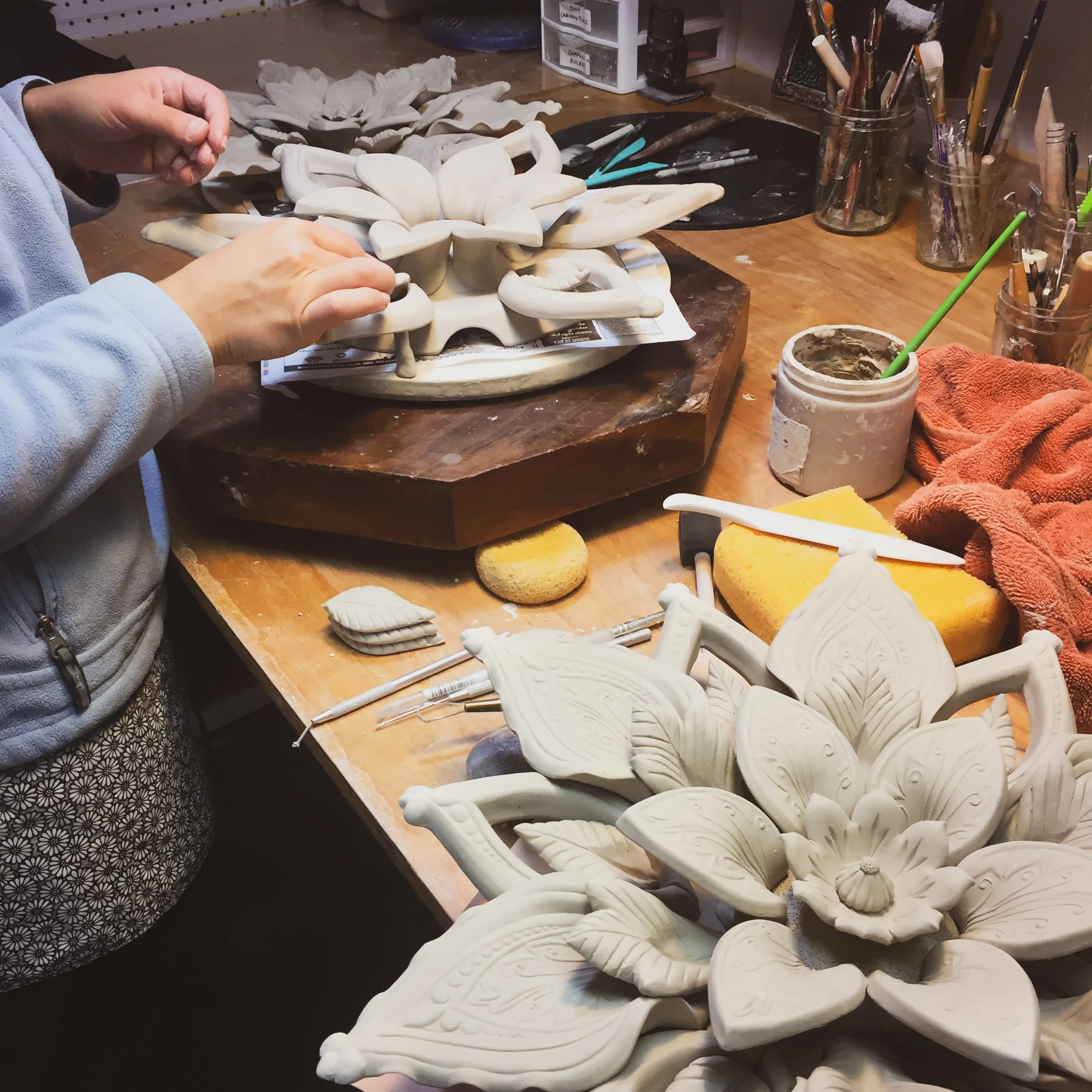 WIP handbuilt porcelain flower on a wooden worktable, with pottery tools, brushes, and containers of clay nearby in an art studio.