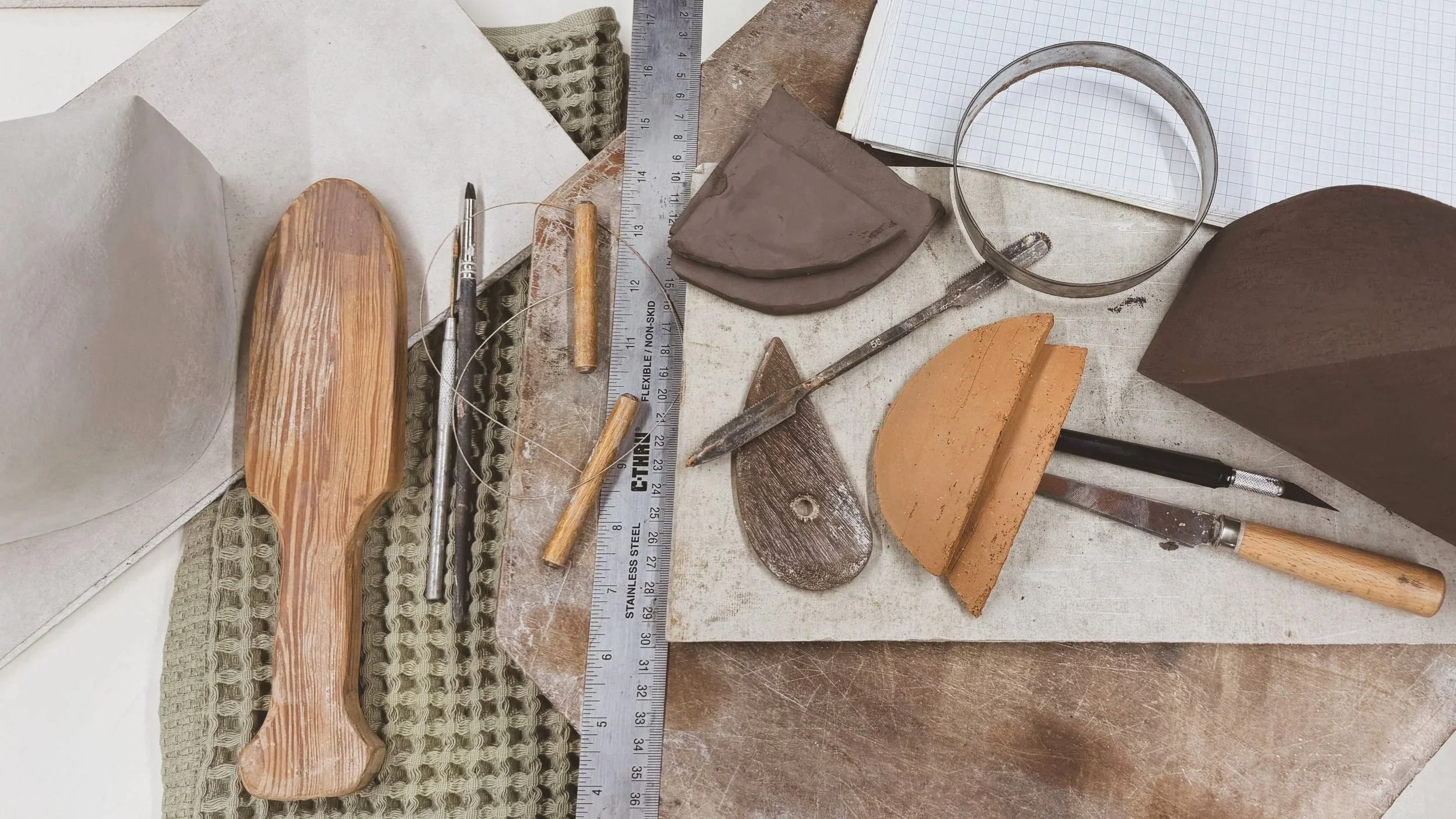 A ceramic workspace with various tools, pieces of clay, a ruler, a cutting tool, and a notepad on a workbench.