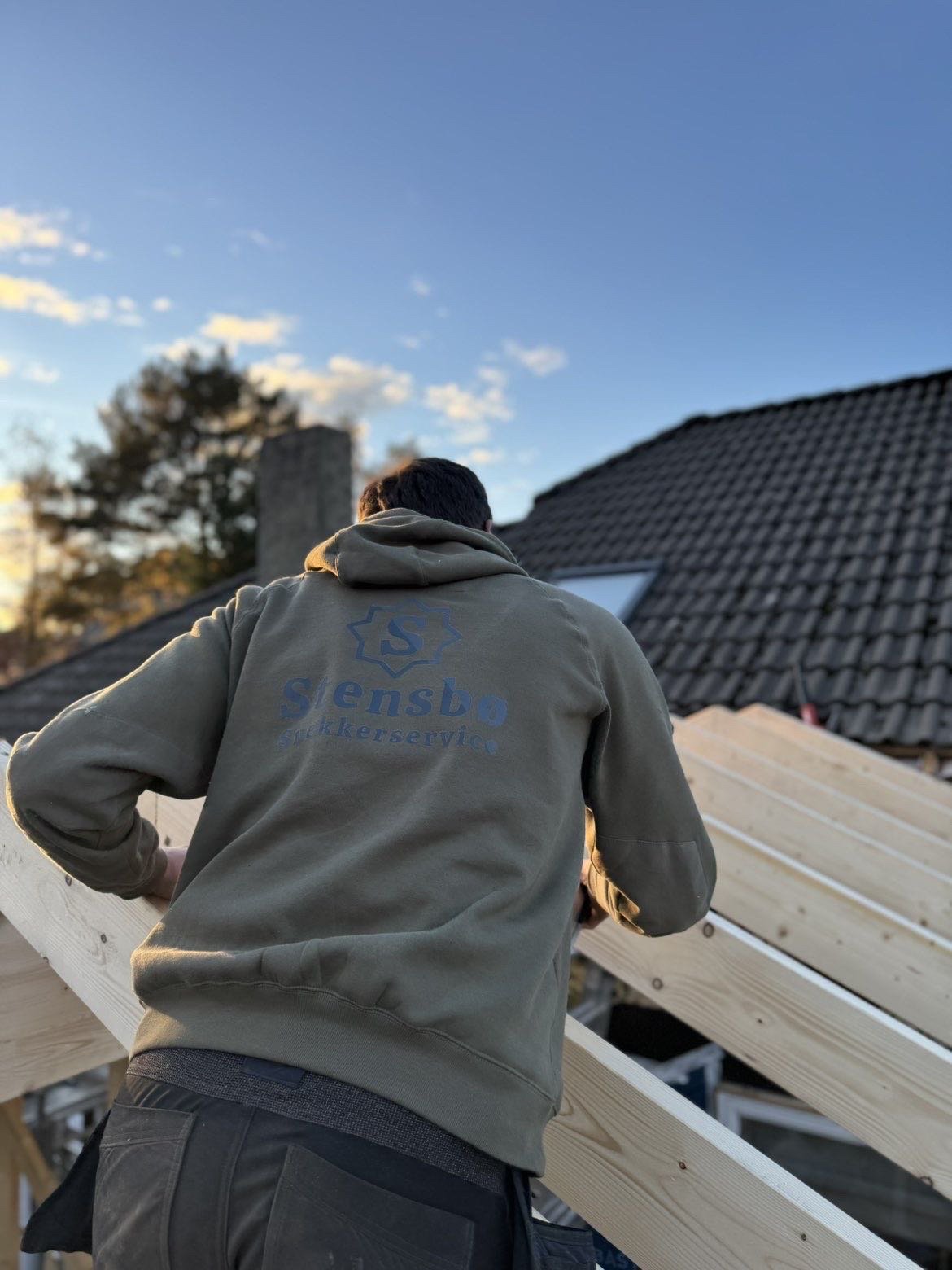 En person står på et tak, der er under konstruksjon, med ryggen mot kameraet. Personen har på seg en grå hettegenser med logo og teksten «Svensbo Rørleggerservice» på ryggen. Solen går ned i bakgrunnen, og det er et klart himmel med noen skyer.