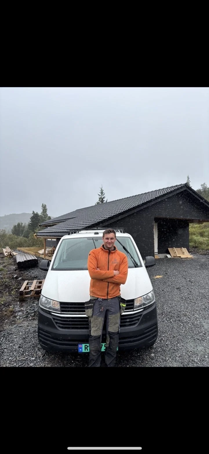 En mann står foran en hvit varebil, iført oransje jakke og bukser, med et svart trehus i bakgrunnen på en grå, regnfull dag.