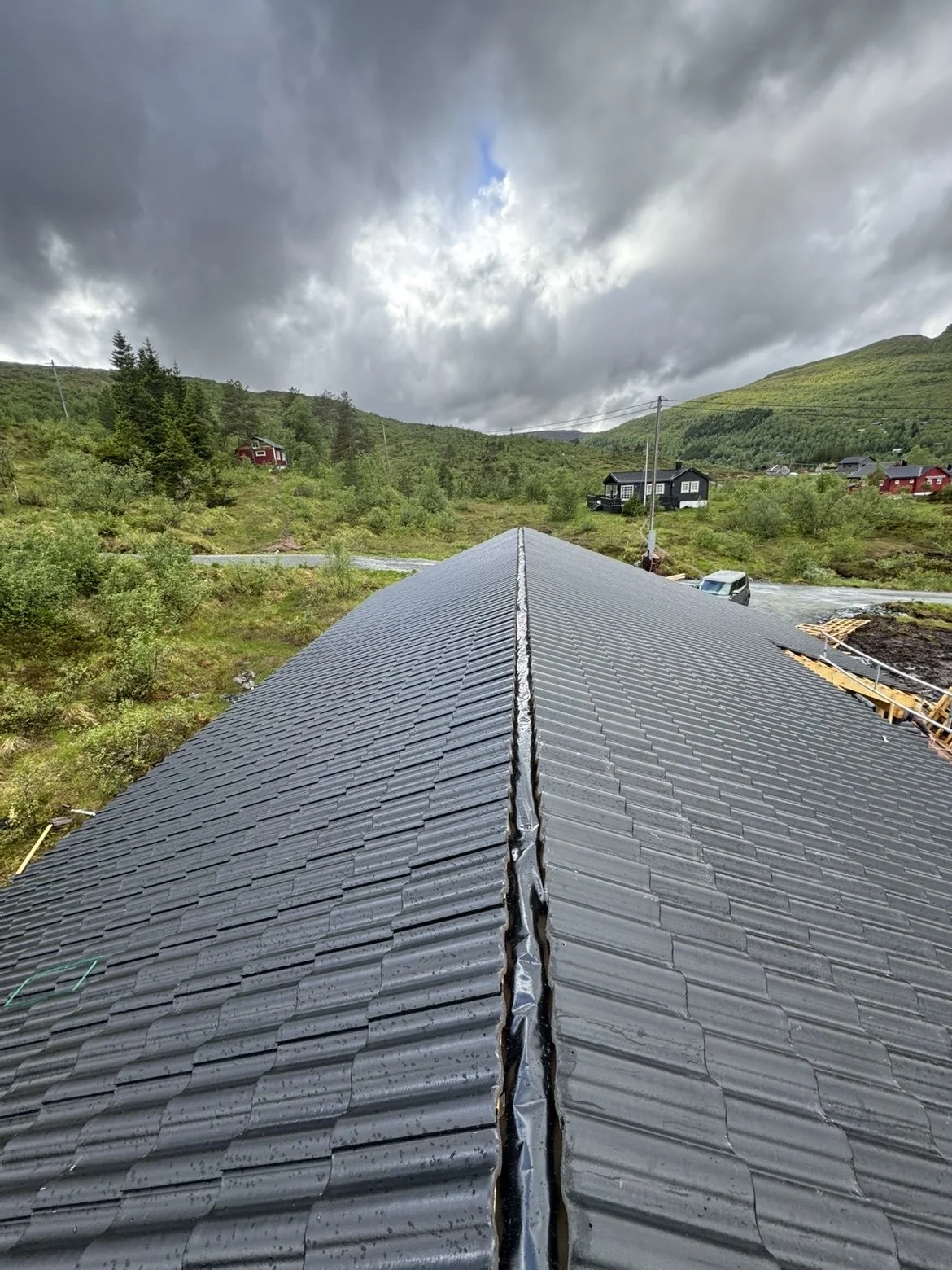 Himmel med skyer over et landskap med fjell, trær og hus. Nærbildet viser taket på en bygning som er under reparasjon.