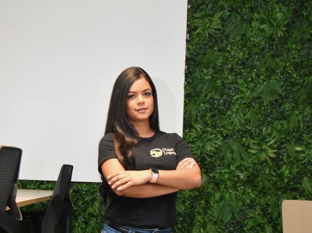 A woman with long dark hair and a watch on her wrist posing with arms crossed, wearing a black T-shirt with a logo, standing in an office with a green wall and a whiteboard.