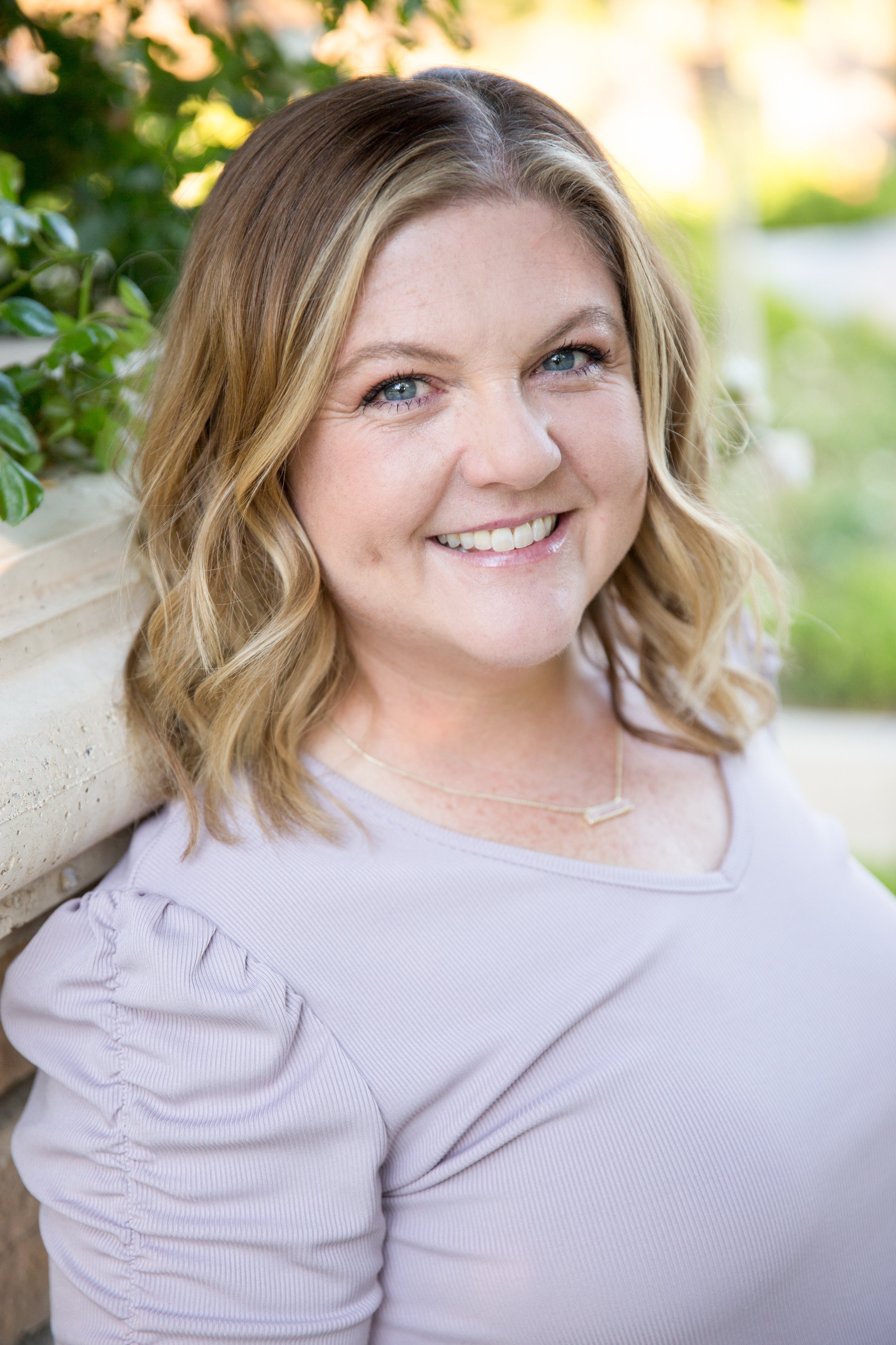 Close-up of a smiling woman with blonde wavy hair, wearing a light purple top, standing outdoors with greenery and a beige wall in the background.