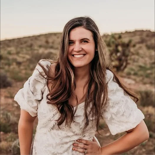 A young woman with long brown hair smiling outdoors in a desert landscape, wearing a cream-colored blouse with puffy sleeves.