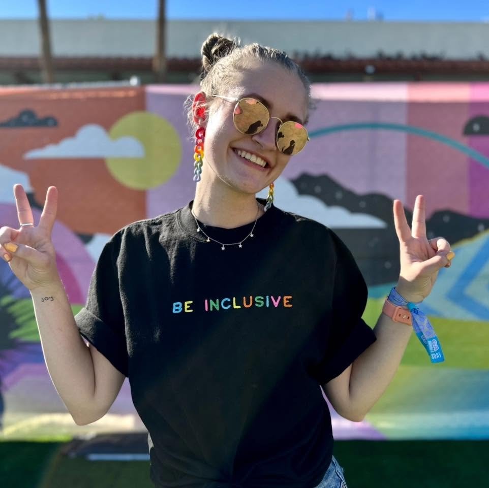 A young woman with rainbow earrings and sunglasses posing with peace signs in front of a colorful mural.