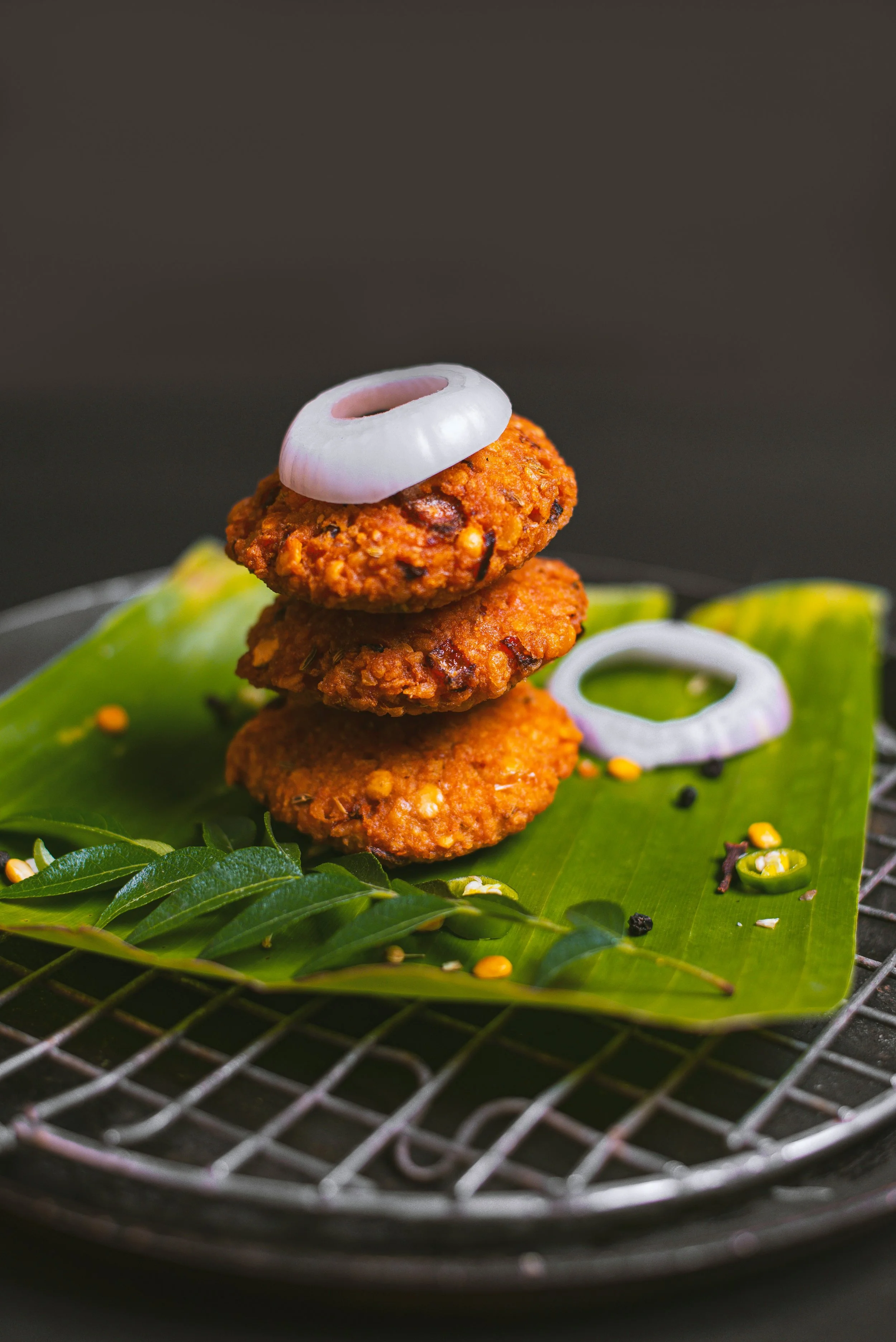 Close-up of three fried vegetarian cutlets stacked on a green leaf, garnished with sliced onions and green chilies on a wire rack.