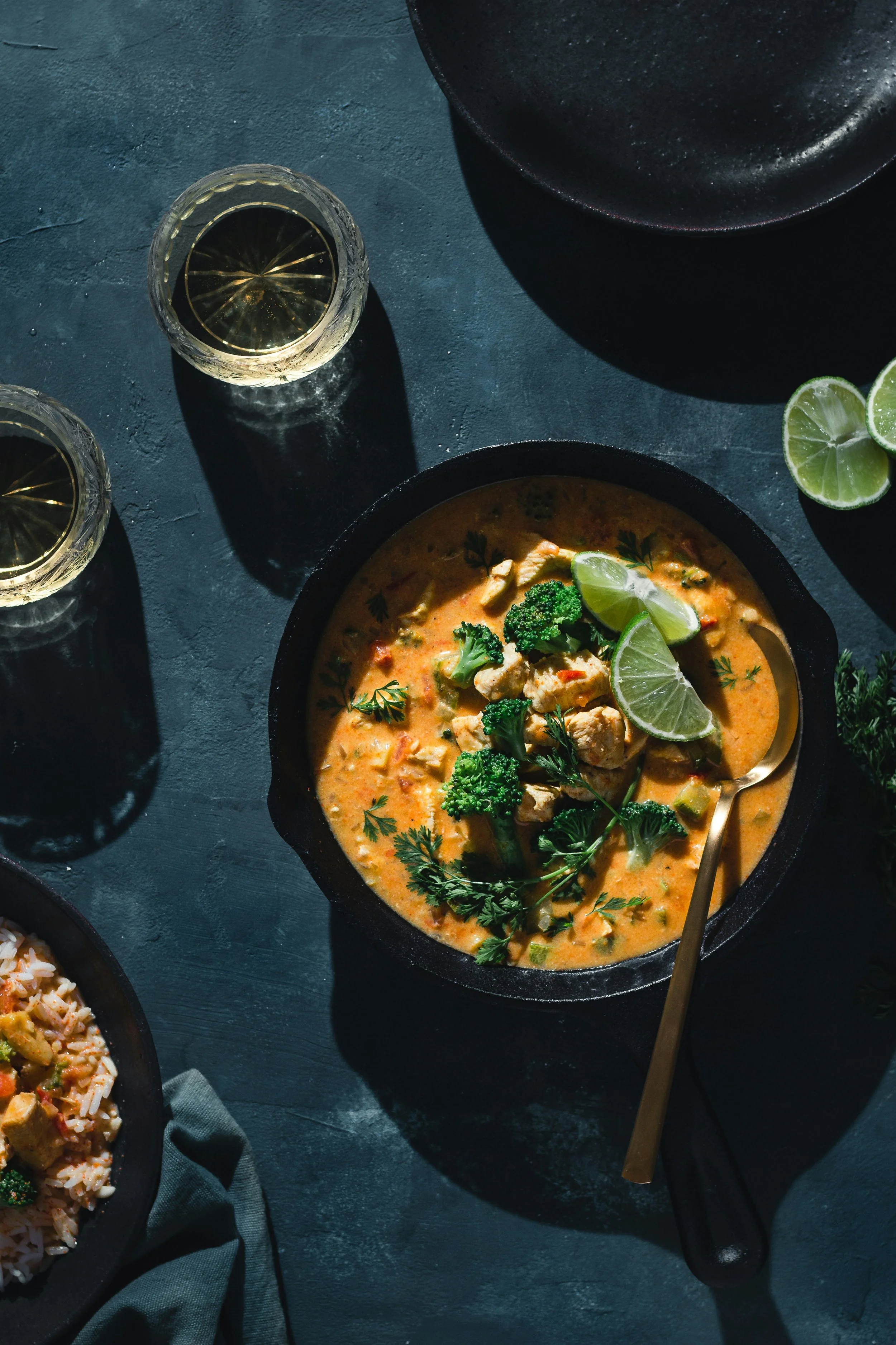 A bowl of chicken and vegetable curry garnished with lime wedges and fresh herbs, alongside glasses of white wine on a dark surface.