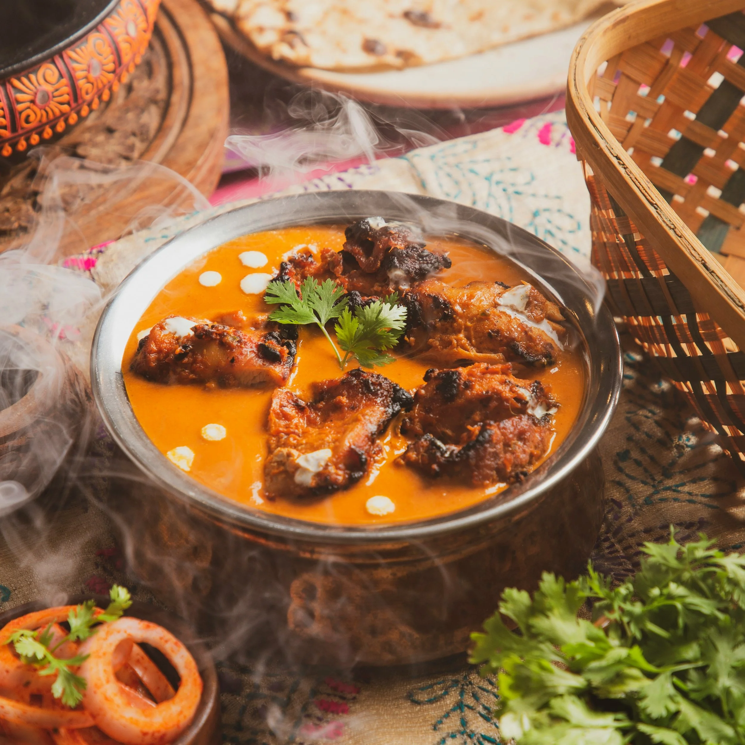 A bowl of orange-colored Indian curry with garnishing of cilantro, served on a decorative cloth with other dishes and woven baskets around.