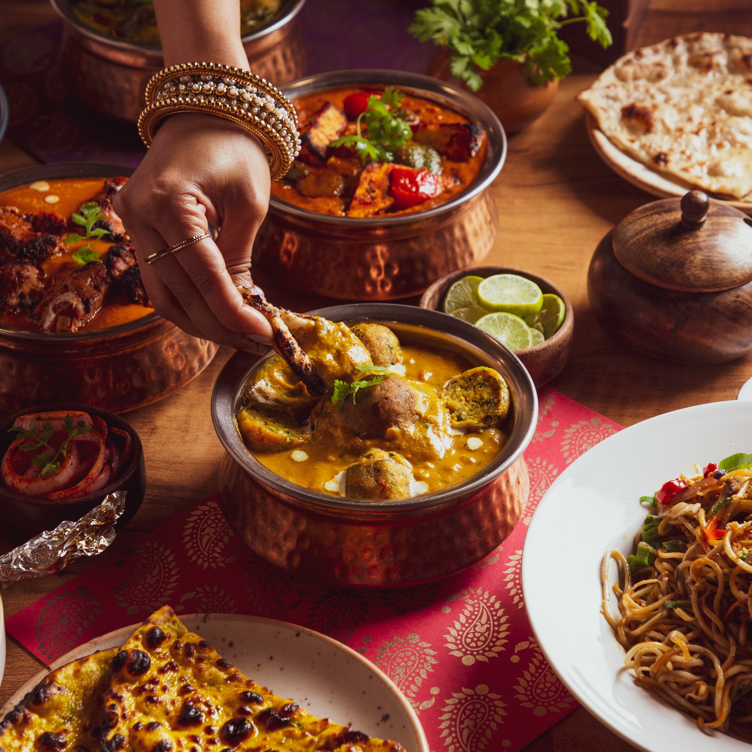 A hand with four layered bangles is reaching for a piece of Indian meat curry. The table is filled with Indian food including a bowl of curry with cilantro, grilled flatbread, lime wedges, and a plate of noodles with vegetables. The table is decorate