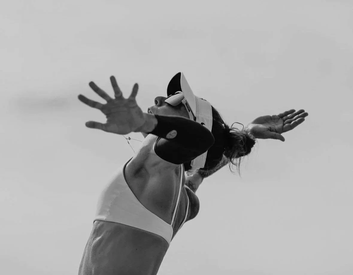 A woman is wearing a helmet and sports attire, reaching out with her arms extended, captured from a low angle against the sky.