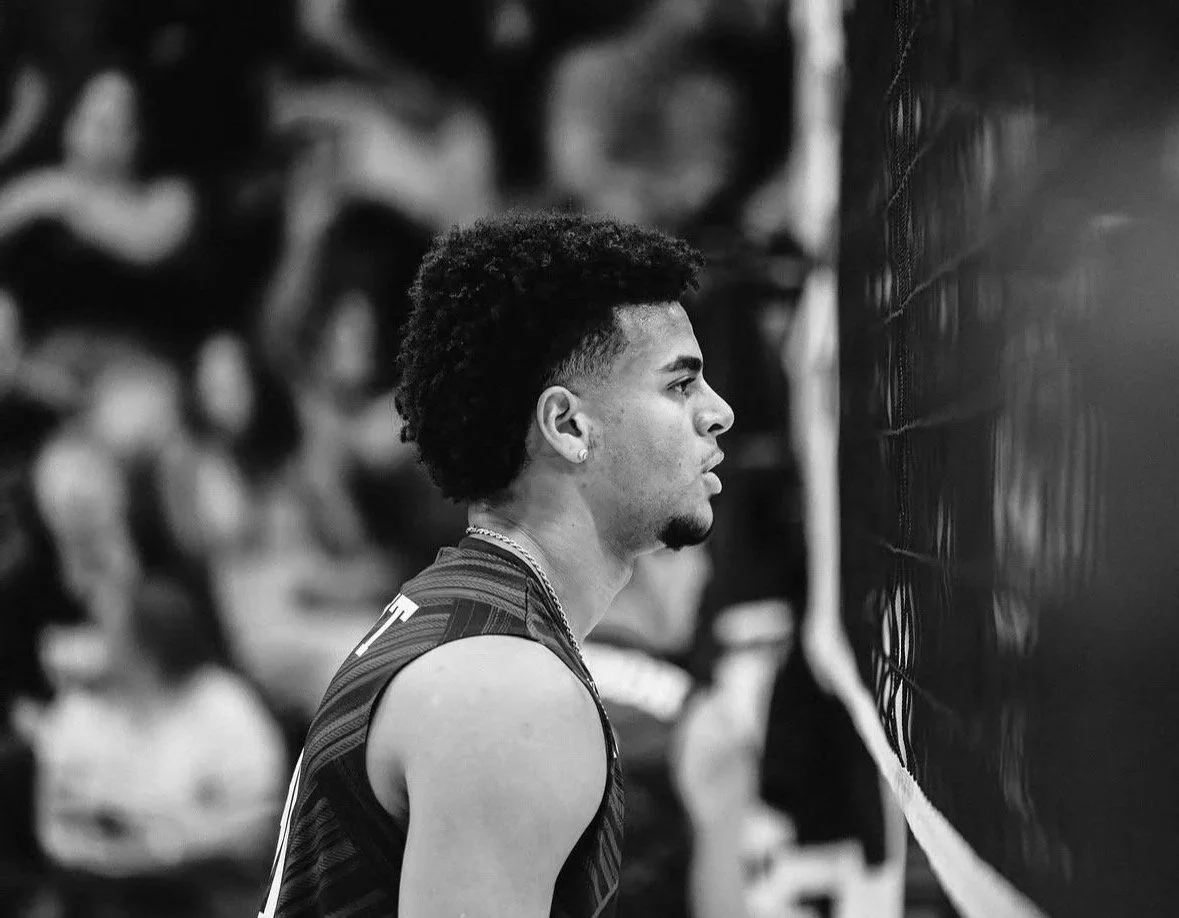 Black and white photo of a male volleyball player in profile, sitting courtside during a match, with a volleyball net in the foreground and blurred spectators in the background.