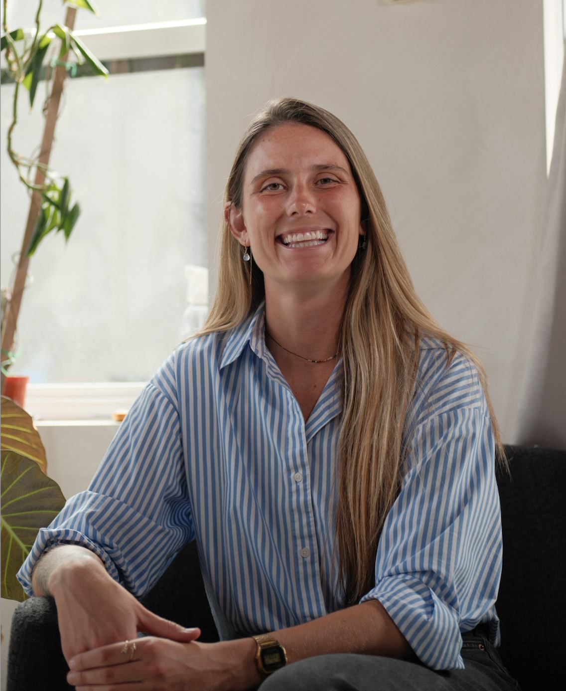 A smiling woman with long blonde hair, wearing a blue and white striped shirt, sitting indoors near a window with plants in the background.