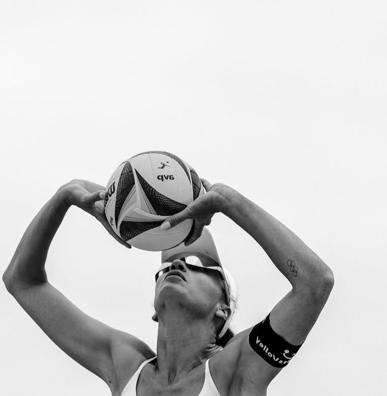A female athlete wearing sunglasses and a sports arm band, holding a volleyball above her head, preparing to serve or hit, with a neutral sky background.