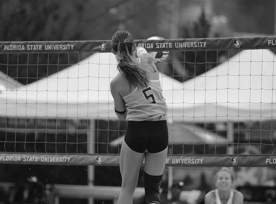 A female volleyball player in a tank top and shorts spikes the ball during a game, with a net labeled 'Florida State University' and other players and spectators in the background.