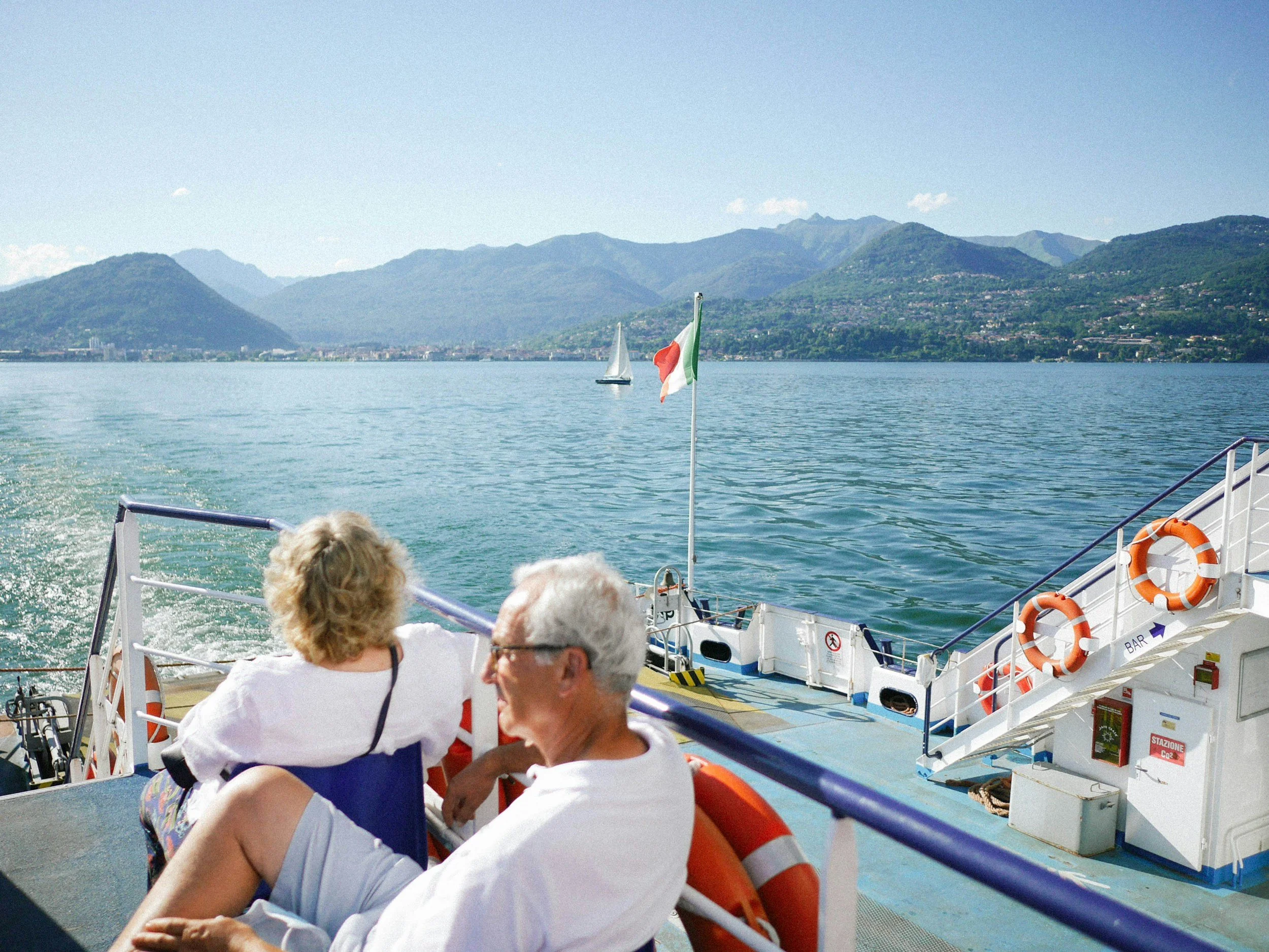 A group of people on a boat sailing across a lake with mountainous terrain in the background. The boat has Italian flags and life preservers on board.