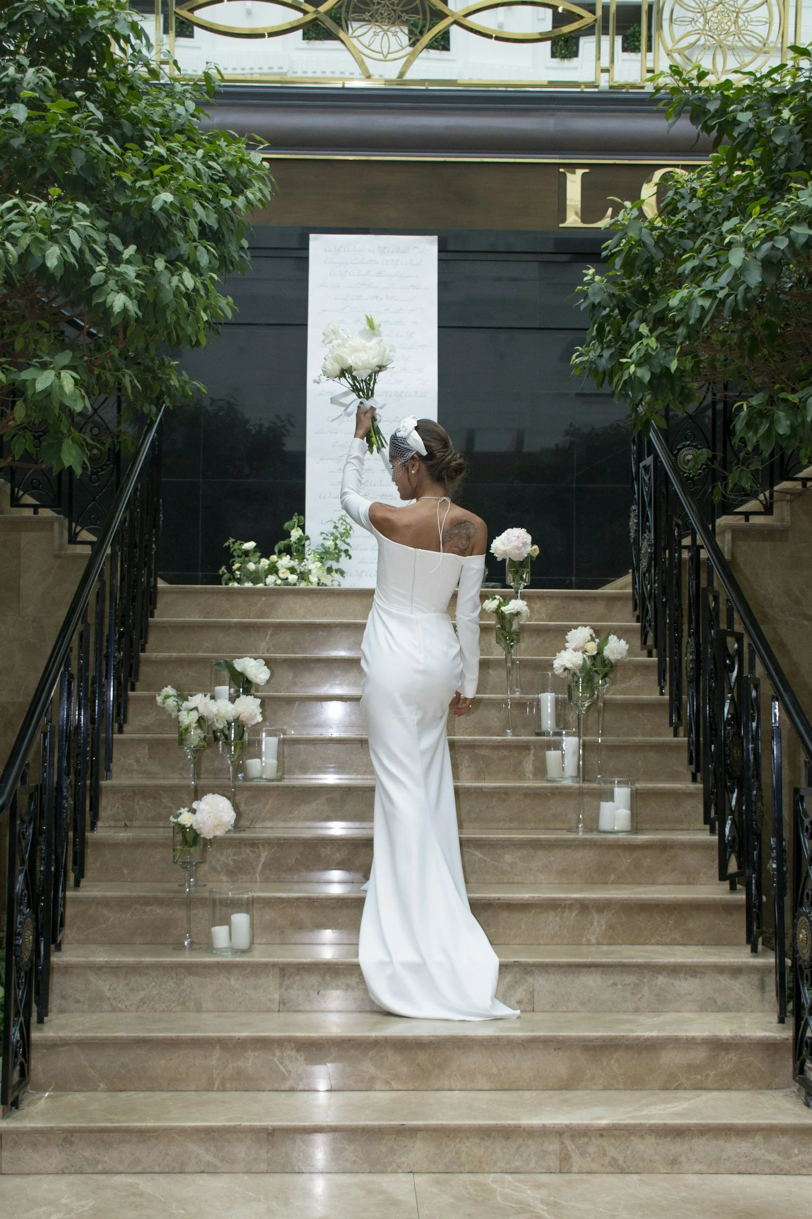 A woman in a white gown holding a bouquet of white flowers standing on a staircase decorated with white flowers and candles. She is facing away, on a staircase flanked by black wrought iron railings, with greenery on either side. In the background, a sign with handwritten text is visible on a vertical board.