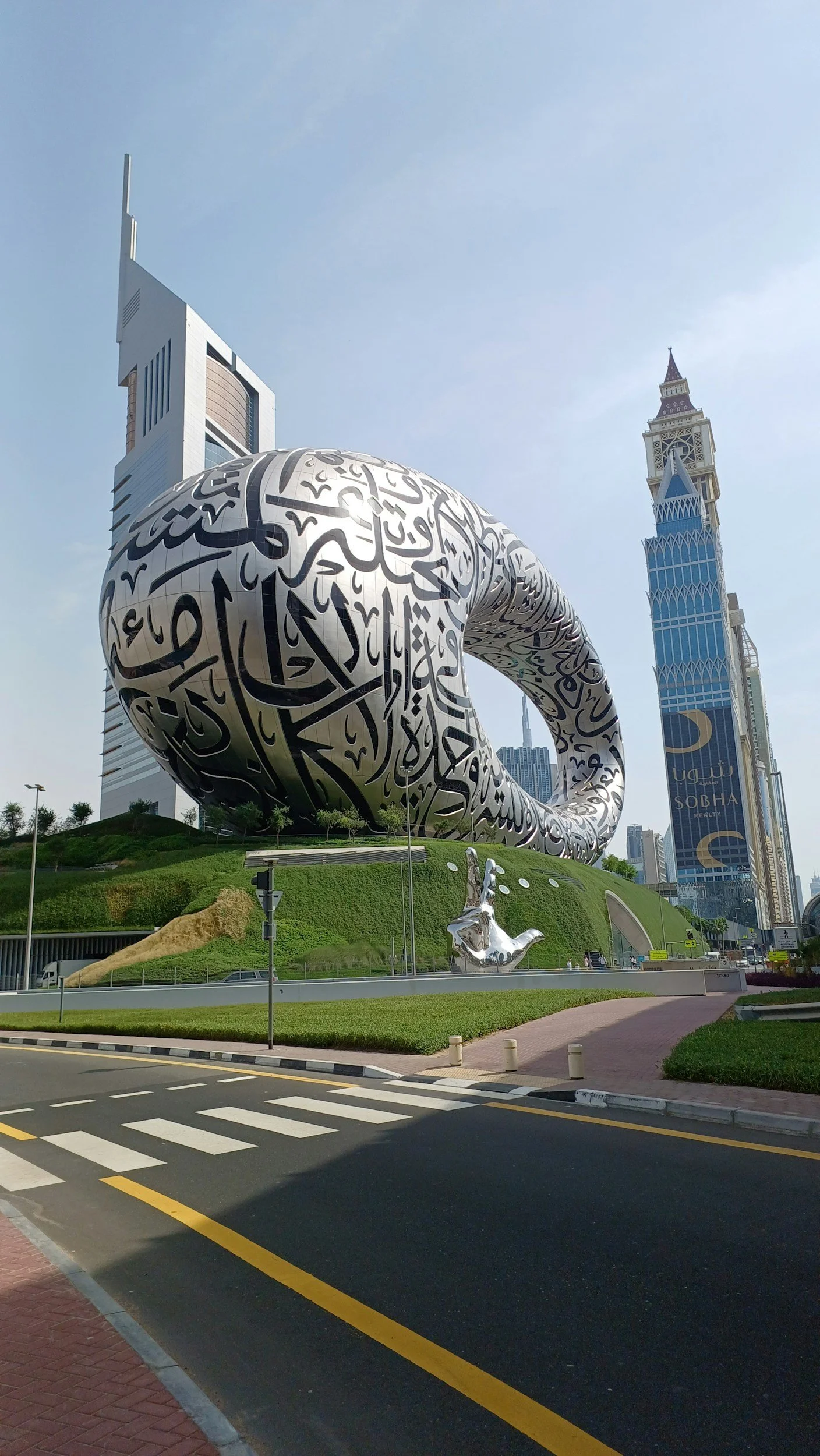 View of the Dubai Frame, a large modern architectural structure with Arabic calligraphy artwork, in Dubai, United Arab Emirates. The background shows a blue sky and skyline buildings, including the clock tower with