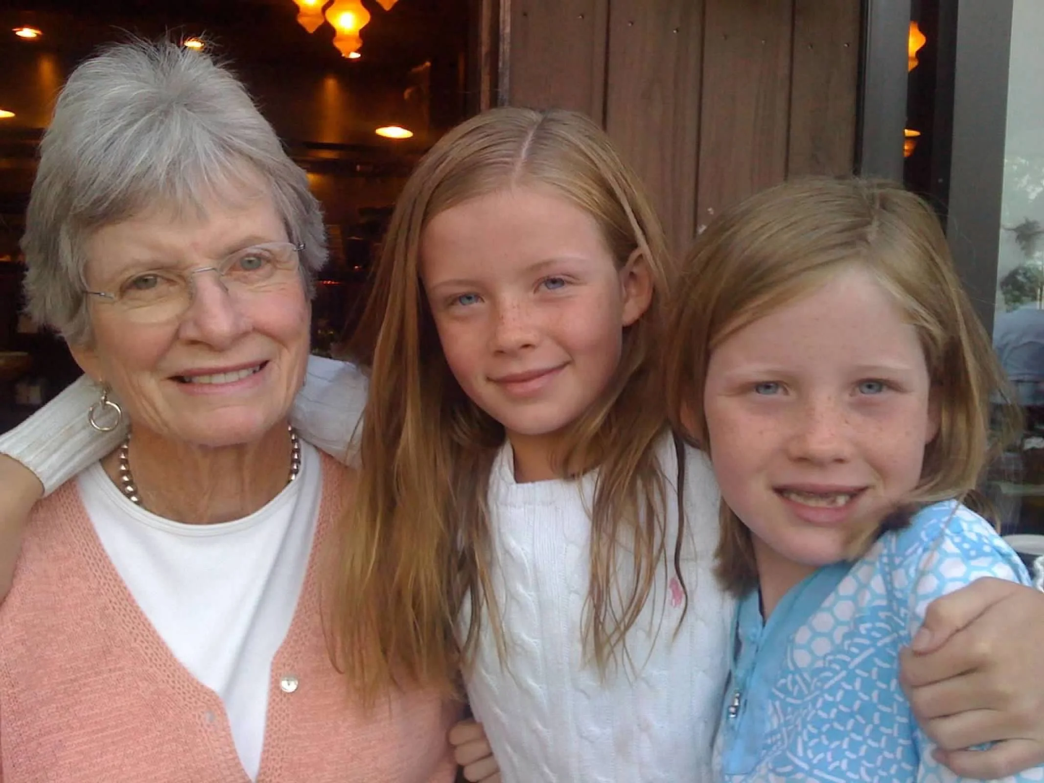 Three women, an elderly woman with gray hair and glasses, and two young girls with red hair, smiling for the camera in a restaurant setting with warm lighting and wooden decor.