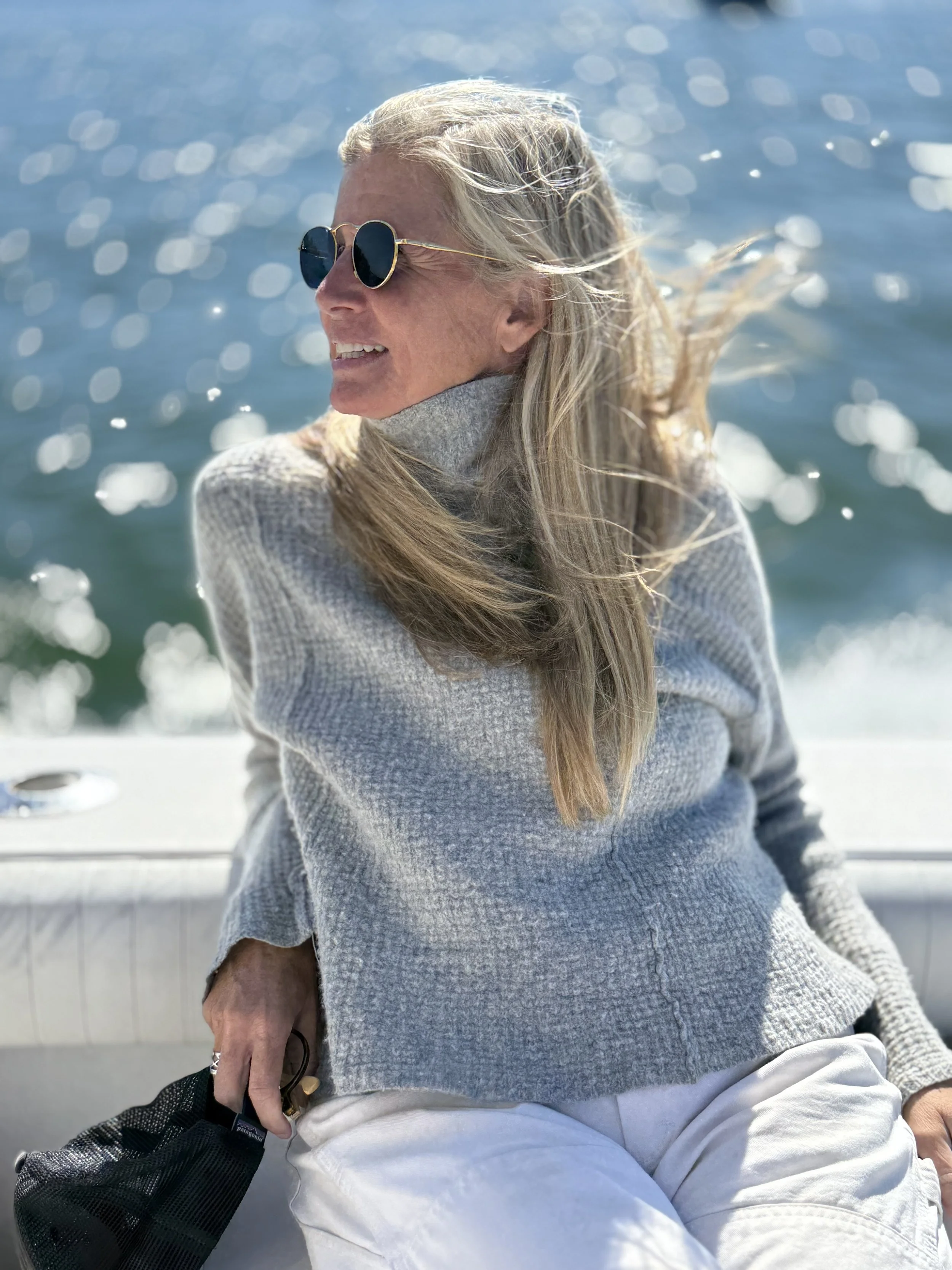 A woman with long blonde hair wearing sunglasses, a gray sweater, and white pants, sitting outdoors by a body of water with sunlight reflecting on the surface, smiling and looking to her left.