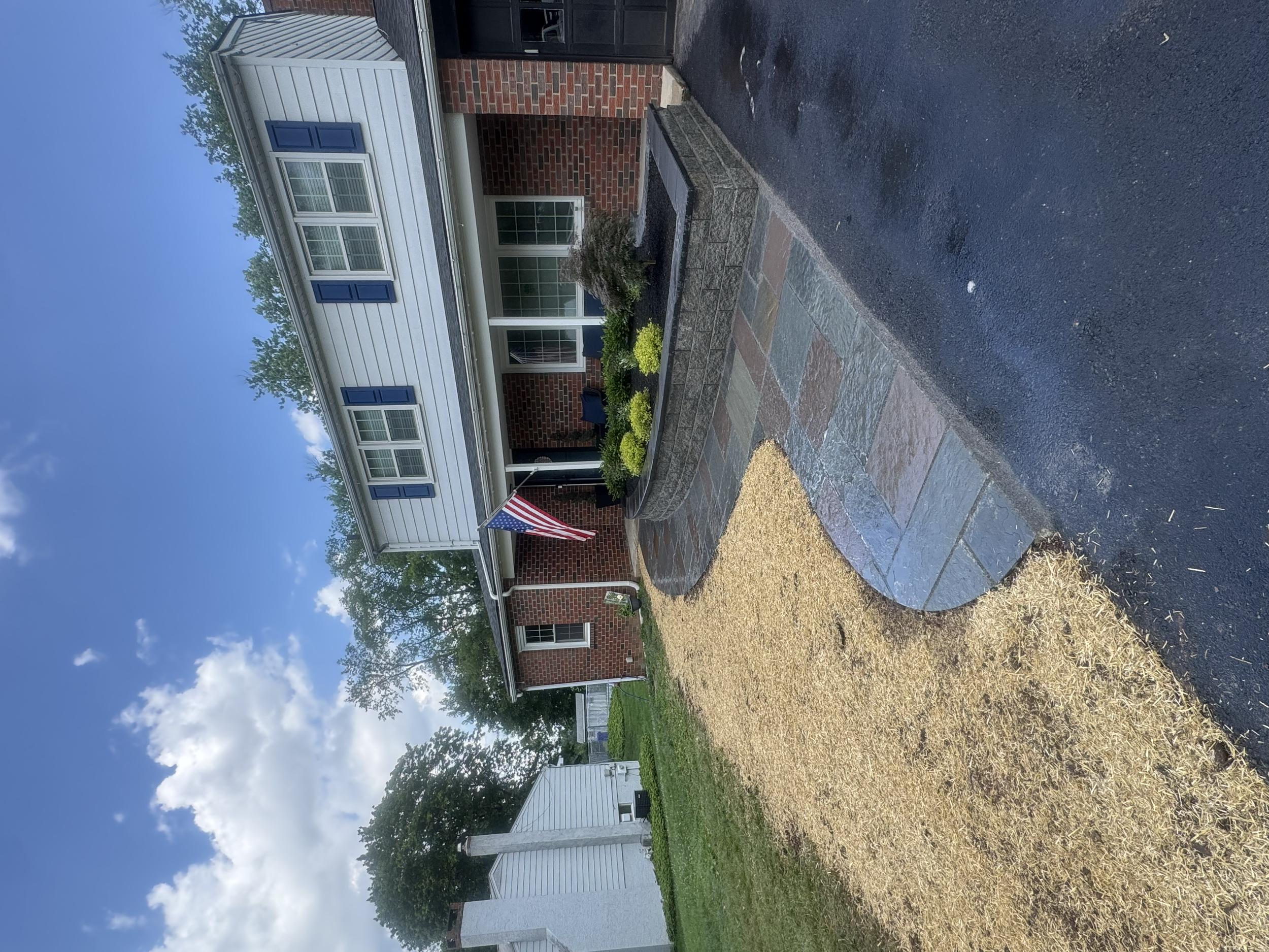 The image shows a two-story house with a brick lower level and a white upper level with blue shutters. In front, there is a small landscaped yard with yellow and green plants, a flagpole with an American flag, and a paved walkway. The sky is partly c