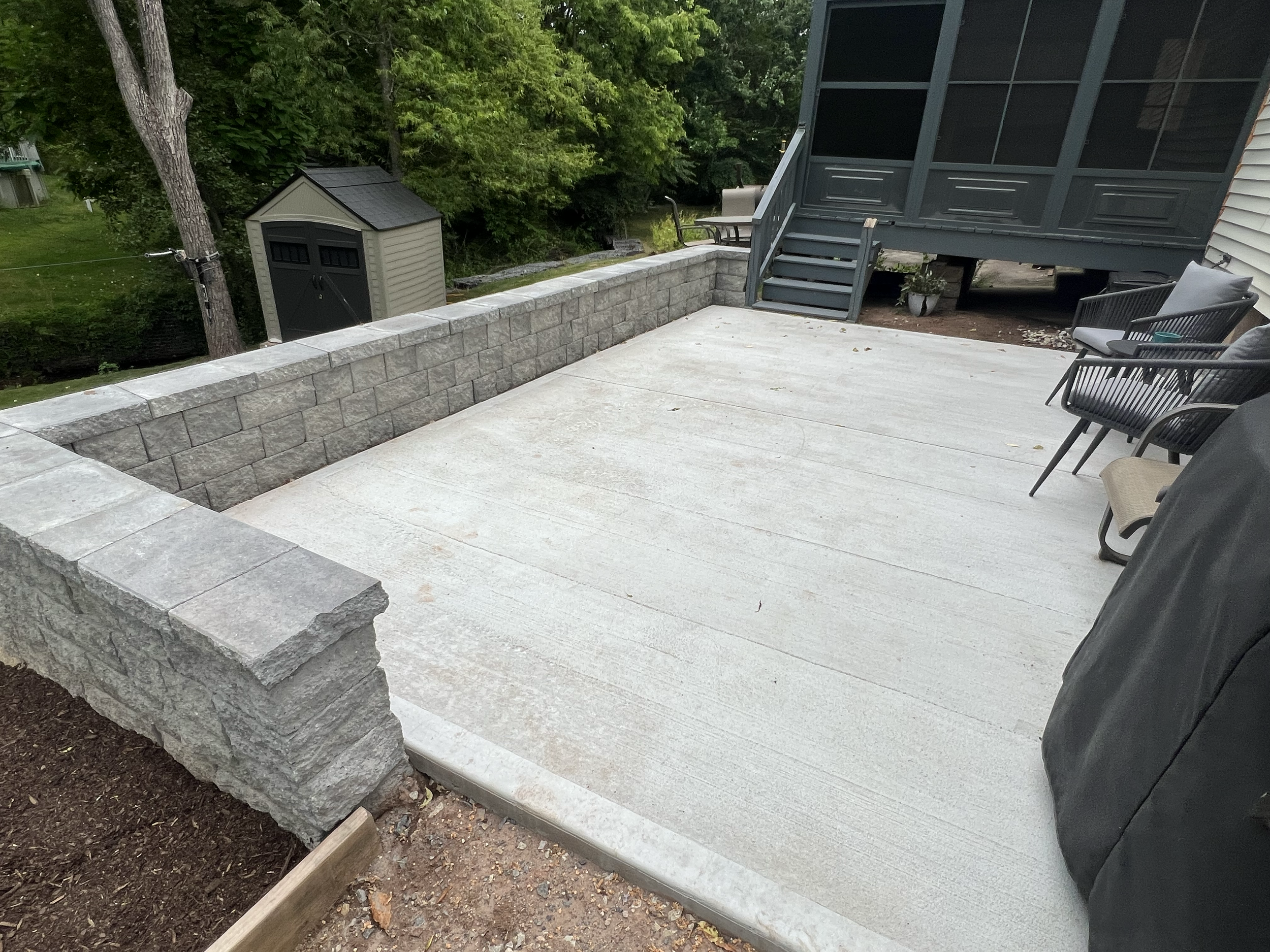 Backyard patio area with a stone retaining wall, concrete flooring, a small storage shed, and a screened porch with stairs, surrounded by trees and greenery.