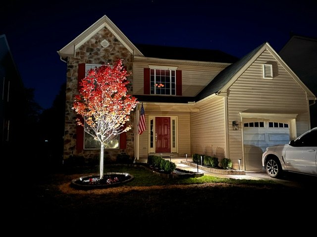 A two-story house at night with lit-up trees in the front yard, American flag, and a white SUV parked outside.