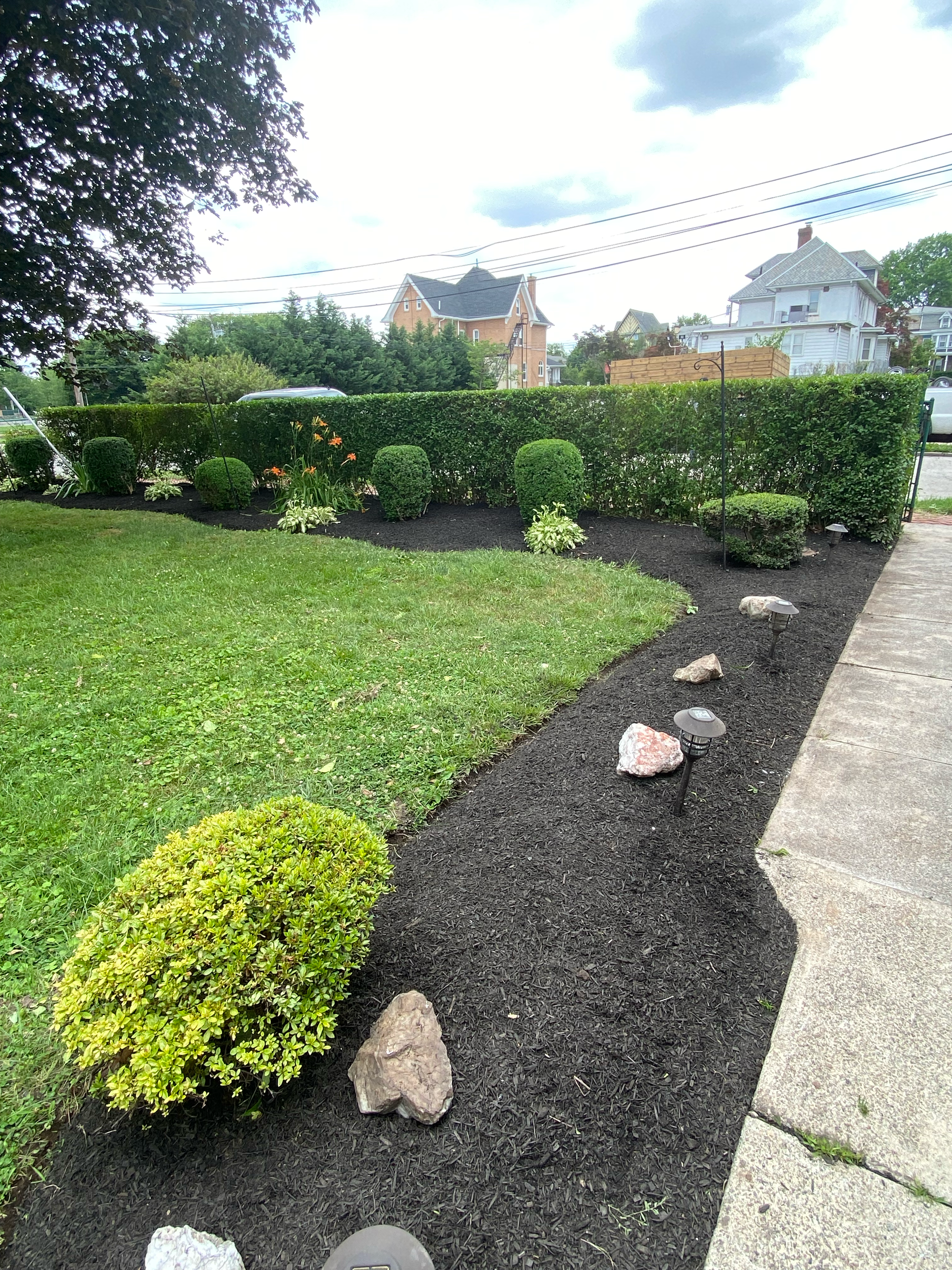 Residential front yard landscaping featuring fresh black mulch, ornamental shrubs, decorative plants, and neatly maintained garden beds along the front of a stone house.
