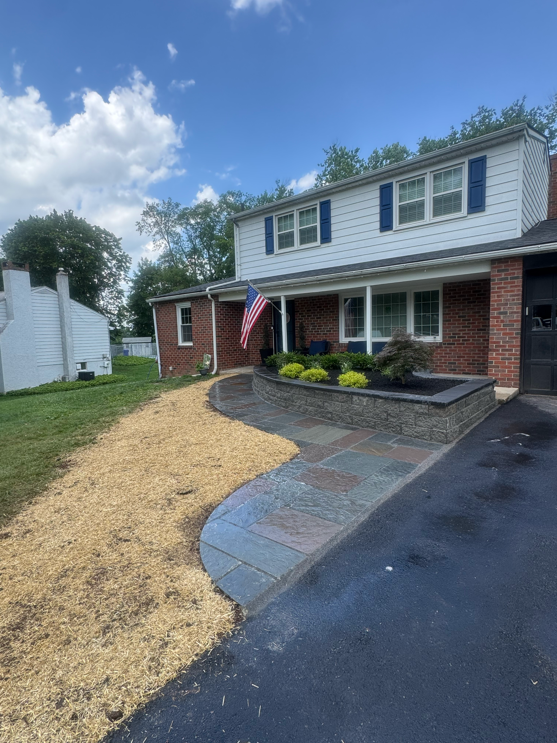 The image shows a two-story house with a brick lower level and a white upper level with blue shutters. In front, there is a small landscaped yard with yellow and green plants, a flagpole with an American flag, and a paved walkway. The sky is partly c