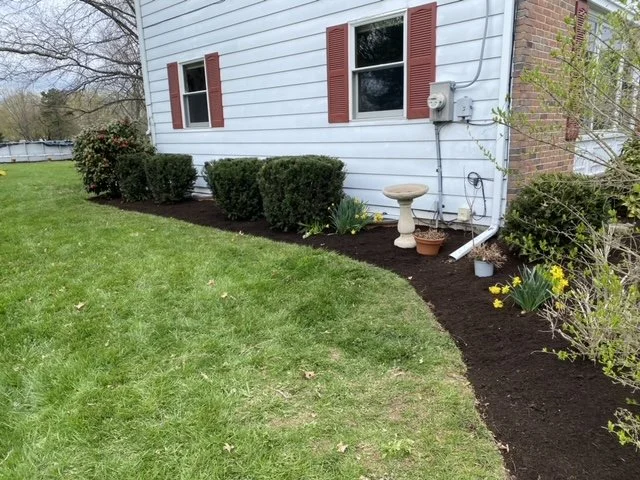 Residential front yard landscaping featuring fresh brown mulch, ornamental shrubs, decorative plants, and neatly maintained garden beds along the front of a stone house.