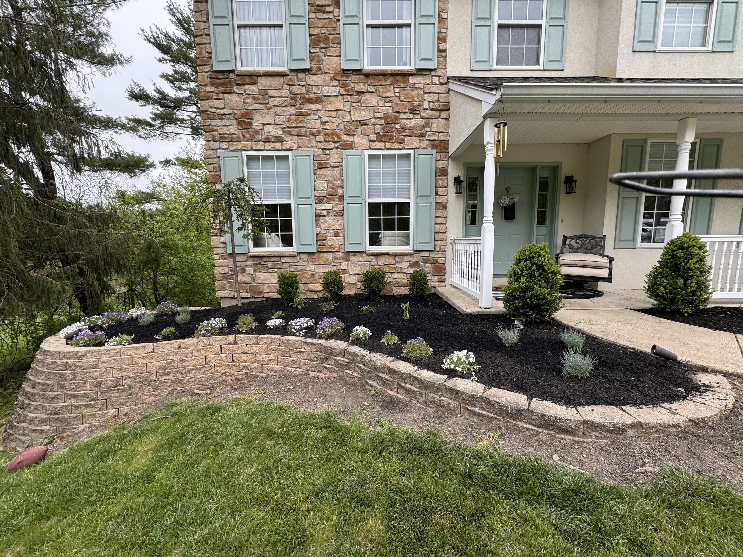 Front yard with a garden bed of small flowering plants, a brick retaining wall, green grass, and a two-story house with stone and stucco exterior, multiple windows with green shutters, a porch with a bench, and a decorative wind chime hanging from th