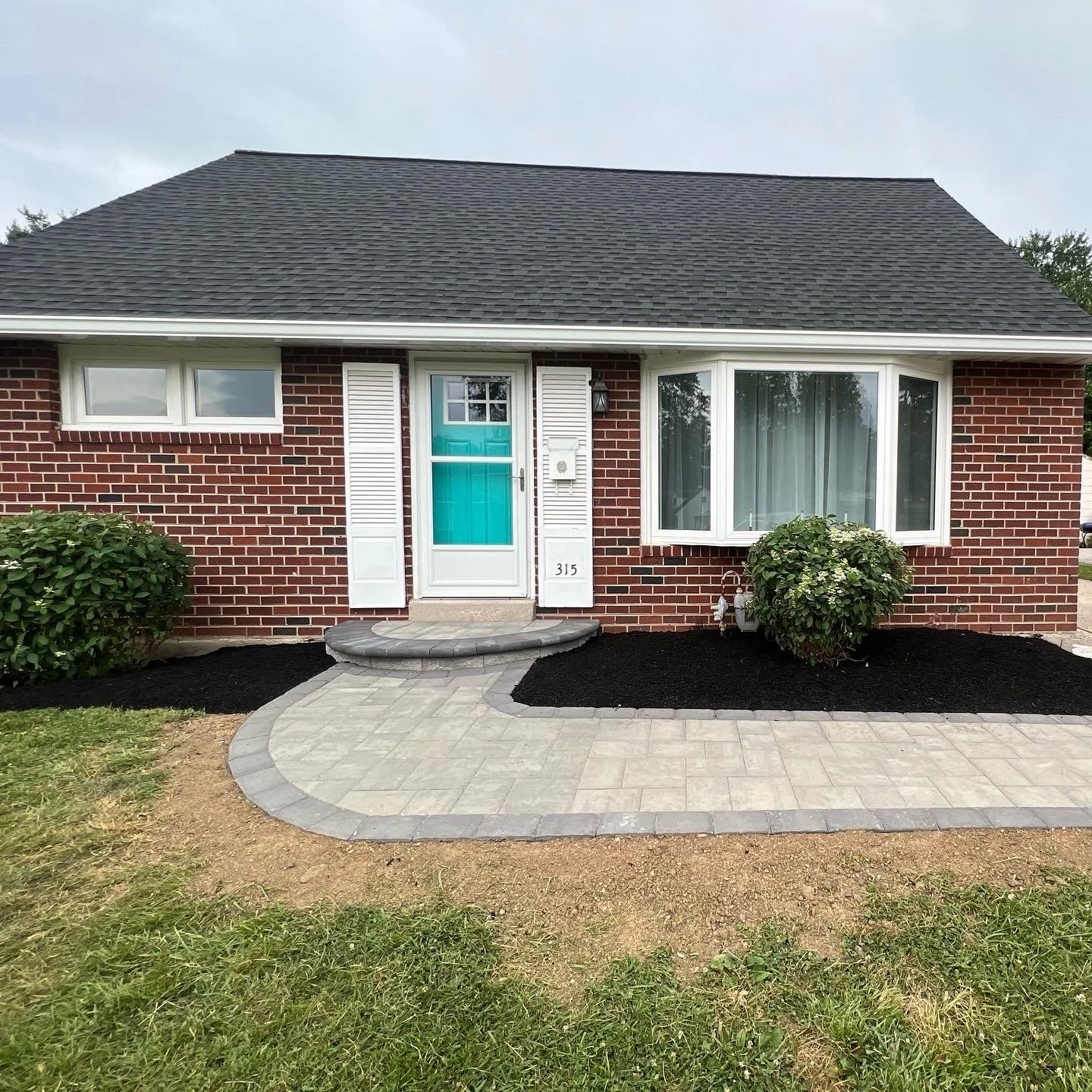 Front of a brick house with a gray shingle roof, white window frames, a turquoise front door, and a curved stone pathway leading to the entrance, surrounded by shrubbery and freshly landscaped black mulch.