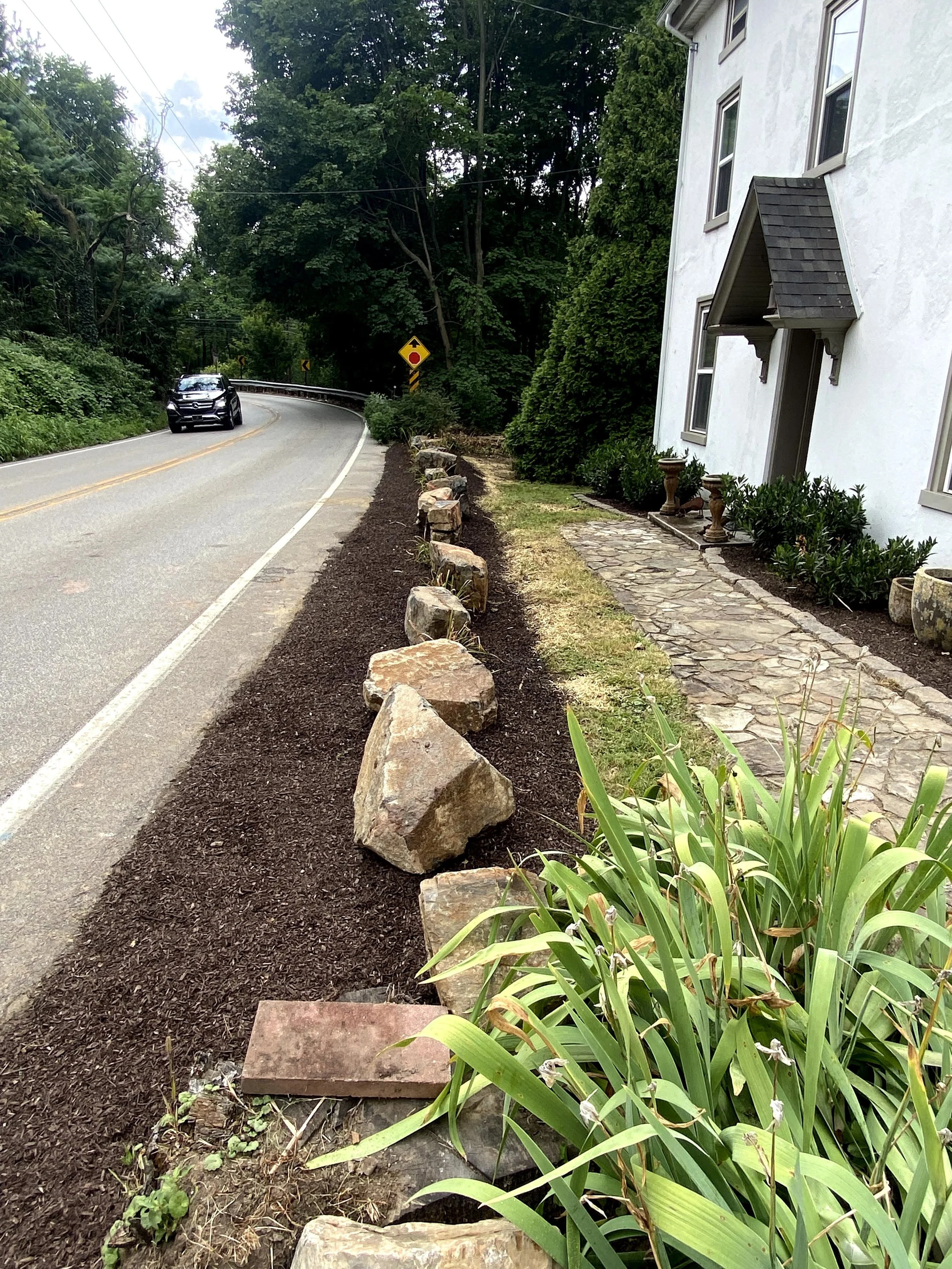 A sidewalk with a row of large rocks separating it from freshly tilled soil with plants. A white building with multiple windows and a small awning is on the right. A black SUV is driving on a winding road with yellow dashed lines ahead, surrounded by